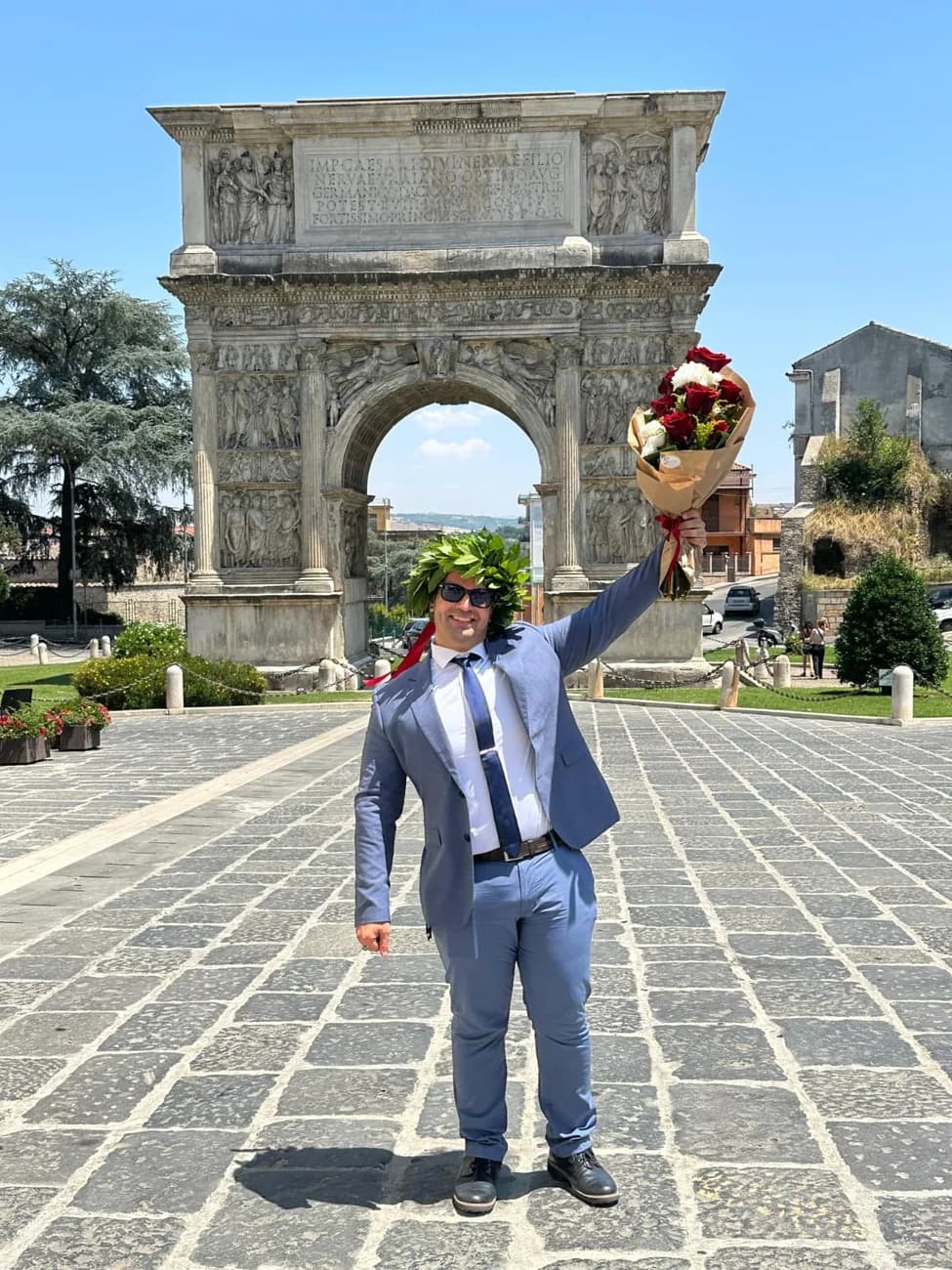 Arman Neyestani in a suit holding a bouquet in front of a stone archway.